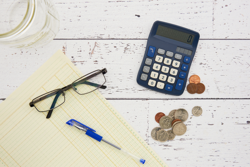 A calculator, coins, reading glasses, and a ledger notepad laid out on a desk for a monthly AI cost breakdown